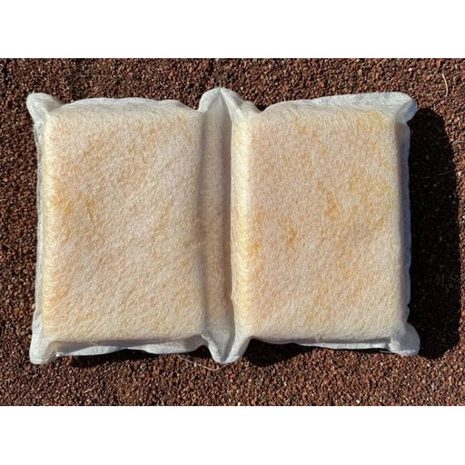 A white sponge pillow with two rectangular sections separated by a central indented seam, resting on a brown gravel surface. The pillow has a fibrous, mesh-like outer texture with a yellowish core visible through the material.