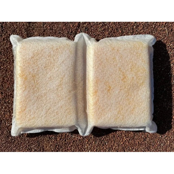 A white sponge pillow with two rectangular sections separated by a central indented seam, resting on a brown gravel surface. The pillow has a fibrous, mesh-like outer texture with a yellowish core visible through the material.