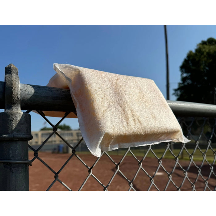 White sponge pillow draped over the top rail of a metal chain-link fence. The pillow's central seam rests on the rail, with one rectangular section hanging on each side. A baseball field and trees are visible in the background under a clear blue sky.