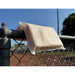 White sponge pillow draped over the top rail of a metal chain-link fence. The pillow's central seam rests on the rail, with one rectangular section hanging on each side. A baseball field and trees are visible in the background under a clear blue sky.