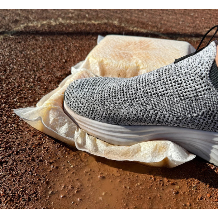 Grey knit sneaker stepping onto a white sponge pillow. The pillow is placed on a muddy, reddish-brown gravel surface, and water is squeezed out from under the shoe as it compresses the sponge. A second sponge pillow sits undisturbed in the background.